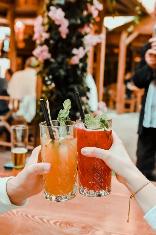 Zwei Hände stoßen mit einem orangen und einem roten Cocktail an